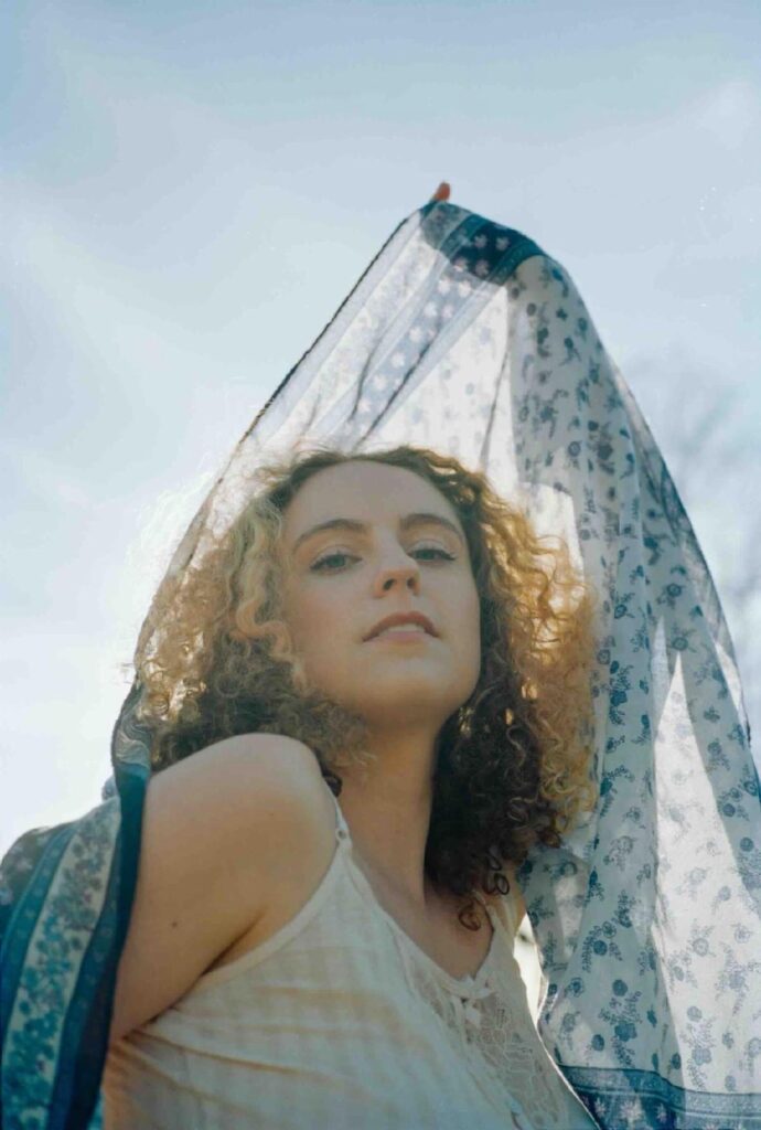 Kinneret stands in a mystical forest scene surrounded by handmade cardboard bird sculptures created by her late grandfather James Grashow. The whimsical, verdant backdrop mirrors the surreal and organic themes of her upcoming album Field Guide
