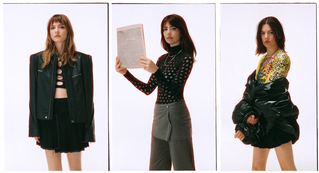 Automatic — Izzy Glaudini, Halle Saxon, and Lola Dompé — pose in moody lighting against a stark background. The trio’s cool, modern aesthetic mirrors the sharp, political edge of their upcoming album Is It Now?.