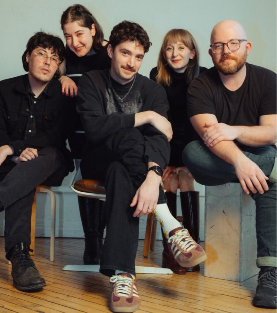Cusp’s Jen Bender sit under stage lights, eyes open, radiating raw emotion bright light shines on them looking smugly.